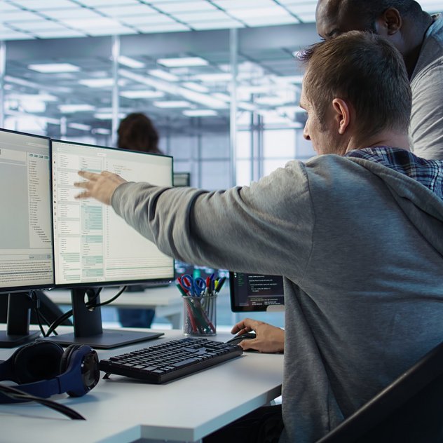 Zwei Entwickler arbeiten eng an mehreren Monitoren; einer zeigt dem anderen den Code im modernen Büro.