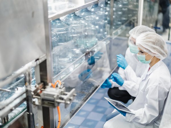 Zwei Fachkräfte in Schutzkleidung prüfen eine Flaschenabfüllanlage hinter Glas, Tablet in der Hand.