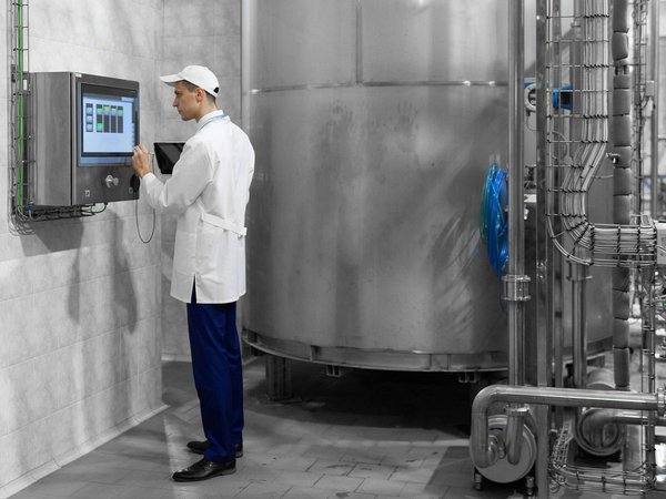 Mitarbeiter im weißen Kittel bedient ein Touchscreen vor einer großen Edelstahlanlage in einer Industriehalle.