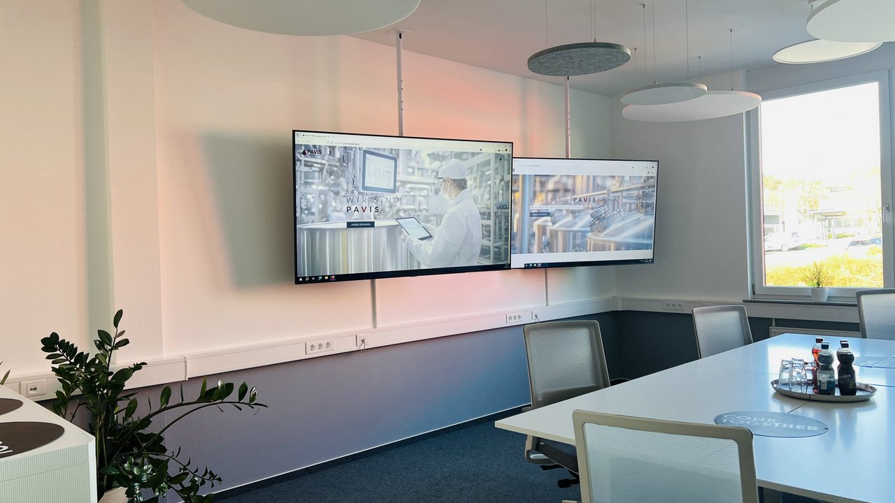 Helles Konferenzzimmer mit zwei Monitoren, die eine Industrieanlage zeigen; Weißer Tisch, Stühle, Pflanzen und Getränke; Fenster.