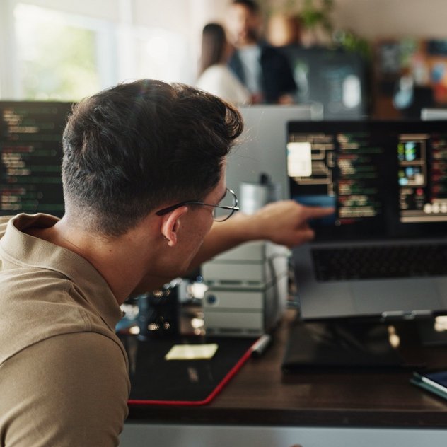 Zwei Entwickler arbeiten an Codescreens am Schreibtisch; einer zeigt dem anderen etwas auf dem Laptop, im Büro mit Monitoren.